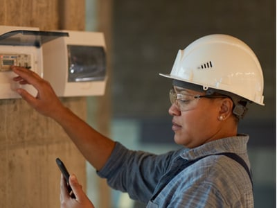 Elektrikerin prüft eine technische Anlage und hält ein Smartphone